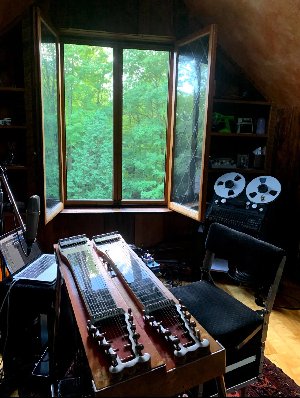 A double-neck steel set up by an open attic window, with a microphone, laptop and tape machine